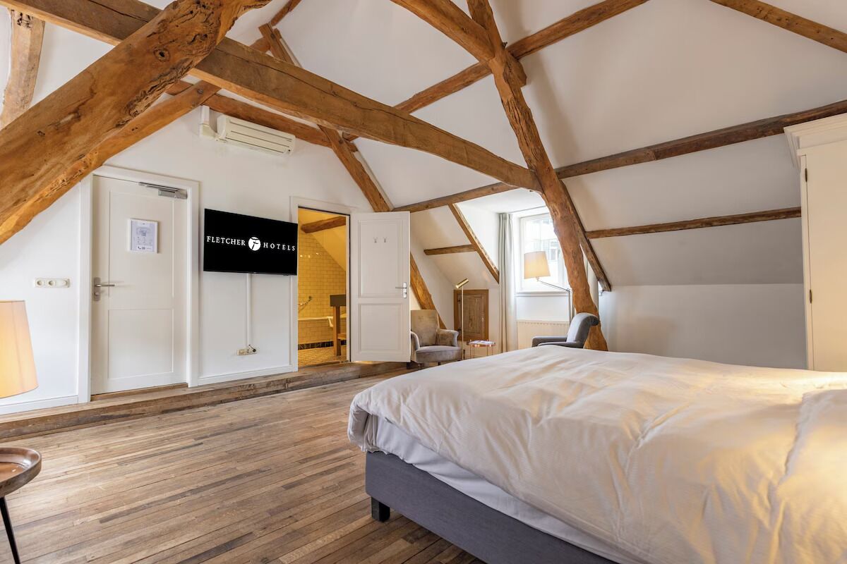 Un dormitorio con techo inclinado, vigas de madera a la vista, una cama, un escritorio y una silla. Se ve un cartel de Fletcher Hotels.