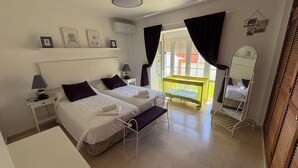 Quarto com duas camas, uma mesa de cabeceira com abajur, uma mesinha com cadeira, janela com cortinas e espelho na parede.