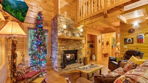 A cozy log cabin interior with a stone fireplace, a Christmas tree, wooden furniture, and a television displaying a mountain view. - Finding Bigfoot 2 Bedroom Cabin (Sevierville)