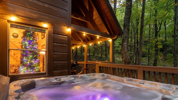 A hot tub with a wooden deck and a Christmas tree inside a cabin surrounded by a forest. - Finding Bigfoot 2 Bedroom Cabin (Sevierville)
