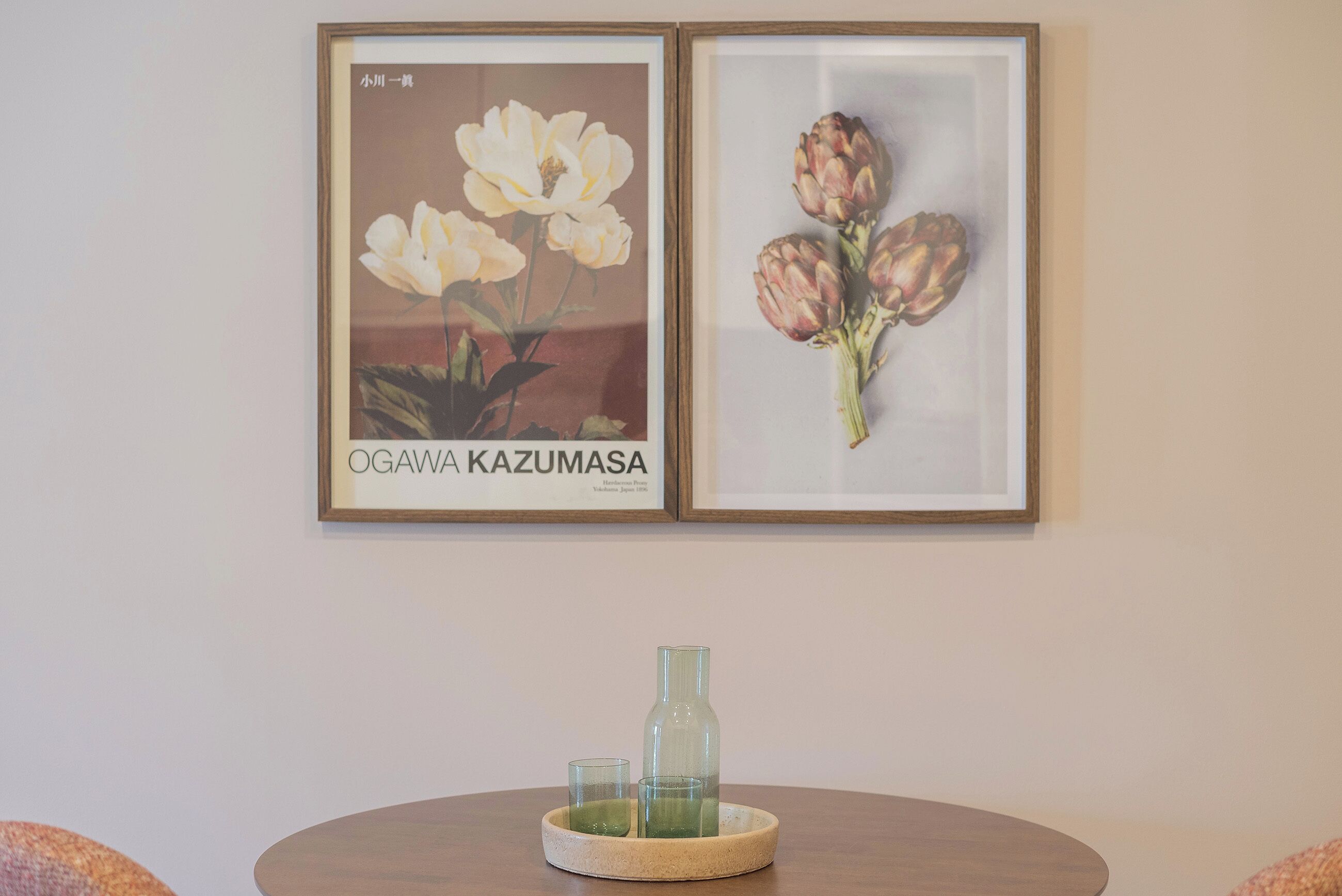 Two framed floral paintings on a wall, a round wooden table with a glass bottle and two glasses, and a textured cushion on the floor.