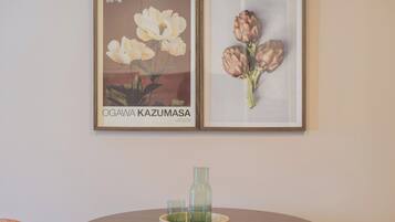 Two framed floral paintings on a wall, a round wooden table with a glass bottle and two glasses, and a textured cushion on the floor.
