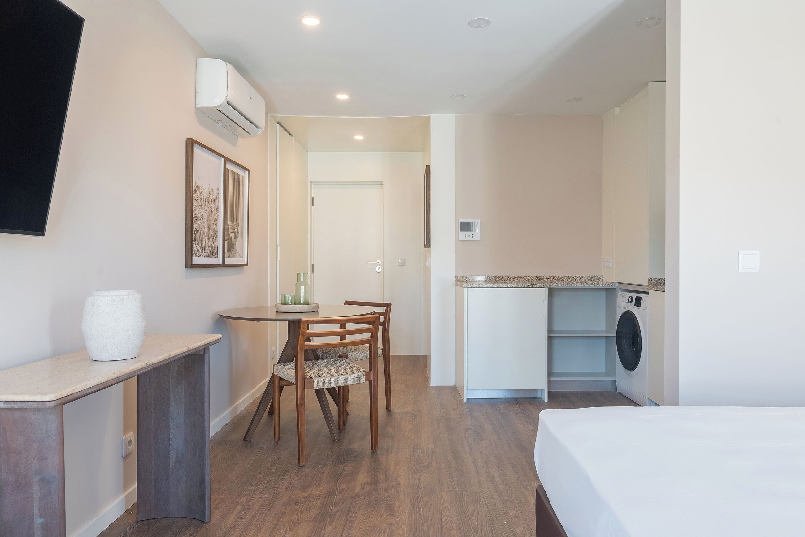 A compact hotel room with a bed, a small dining table with chairs, a kitchenette with a washing machine, and a mounted air conditioner.