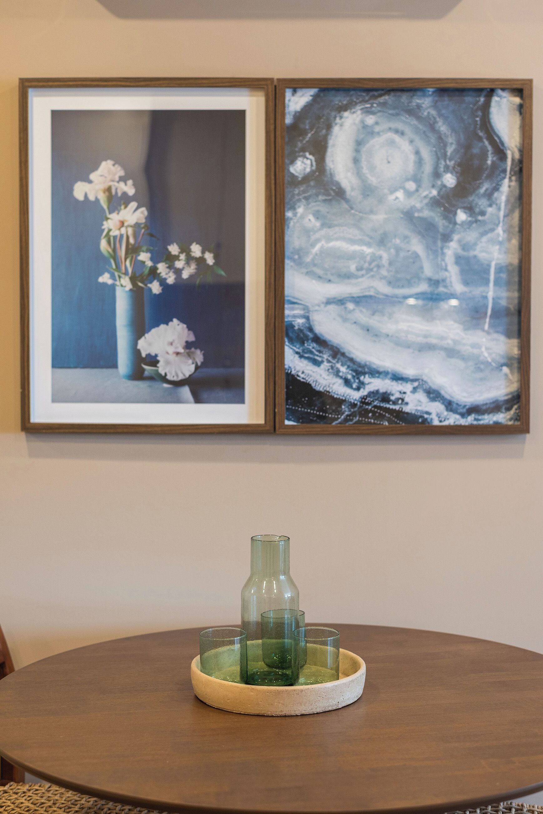 A framed artwork of a flower arrangement on a wall, a glass vase on a round table, and a framed abstract painting.