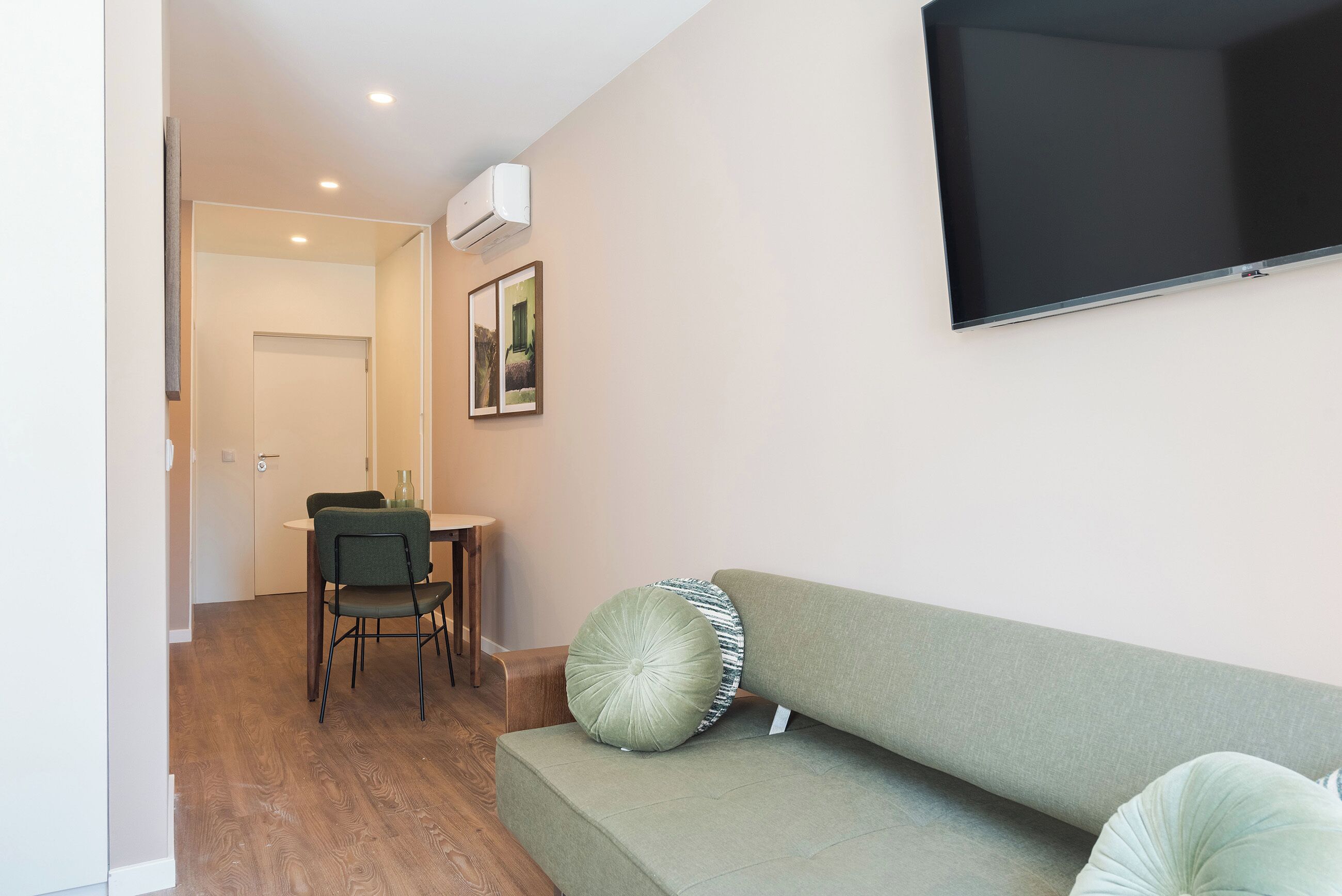 A living room with a green sofa, a flat-screen TV, a dining table with chairs, and an air conditioning unit on the wall.