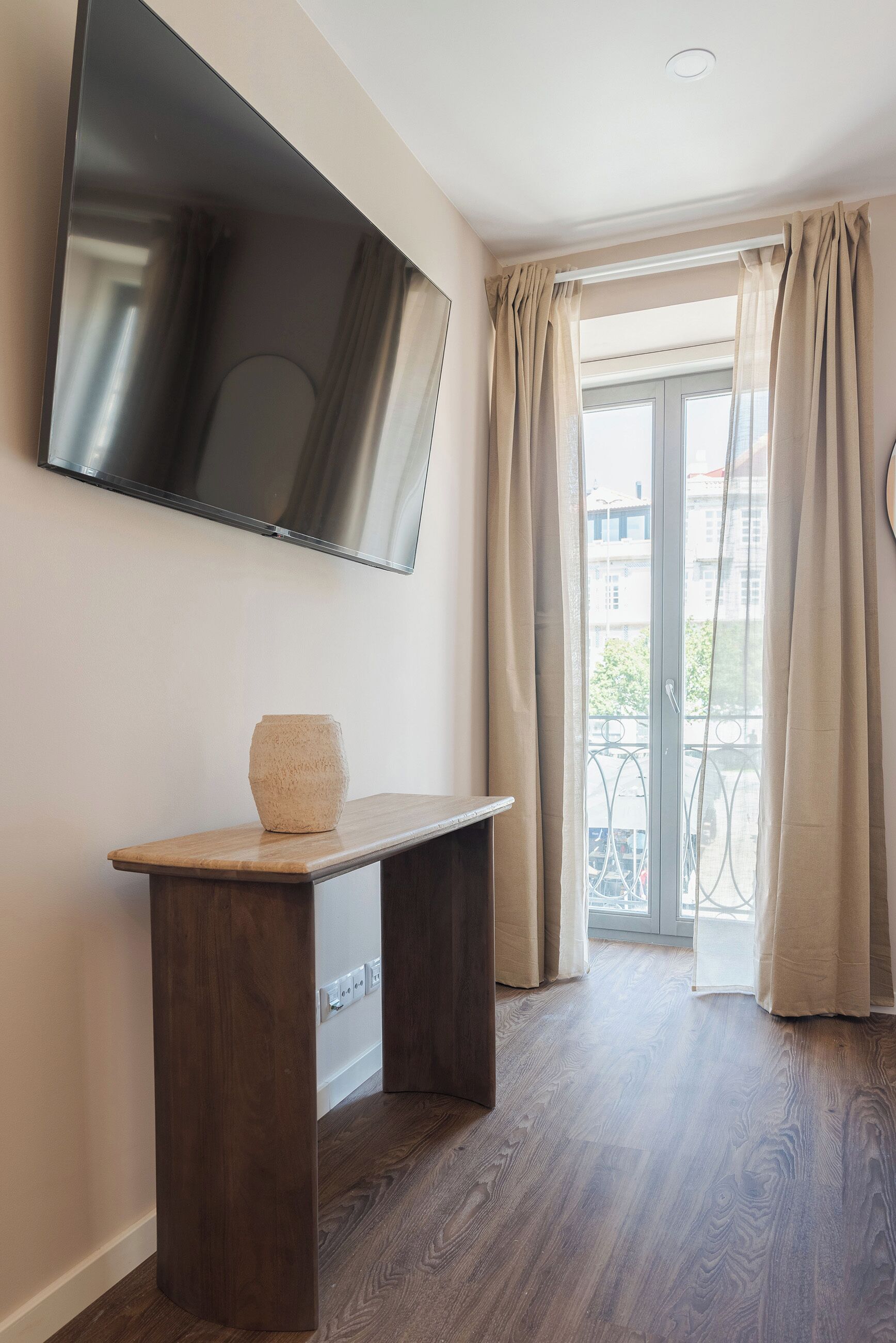 A room with a wooden floor, a flat-screen TV mounted on the wall, a small wooden table with a vase, and a sliding glass door leading to an outdoor balcony.