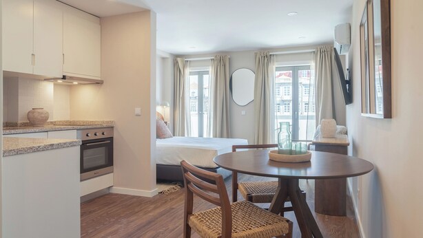 Una camera d'albergo compatta con angolo cottura, un letto, un'area pranzo e ampie finestre con tende.