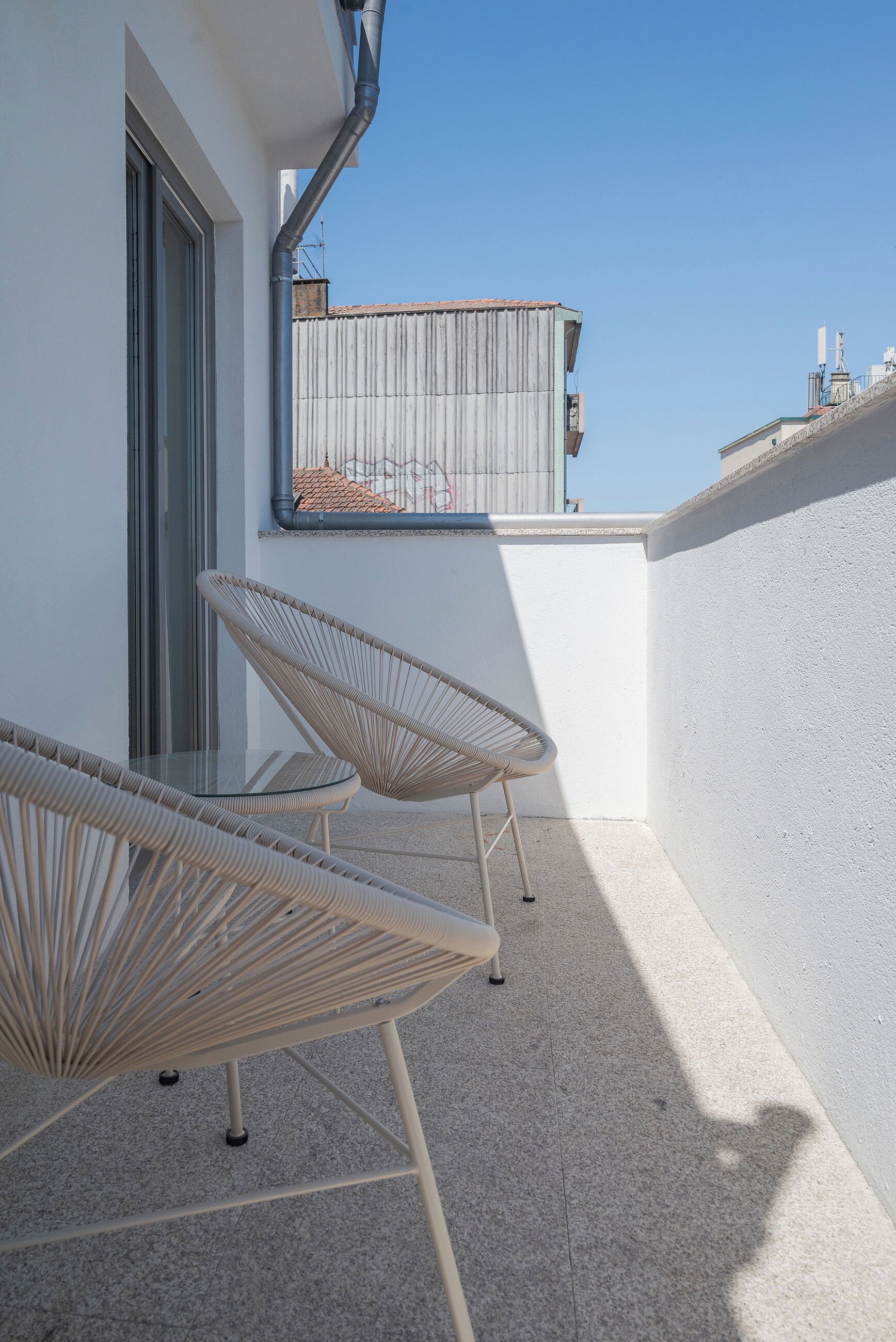 En balkon med to fletstole og et glasbord, hvide vægge og en klar blå himmel.