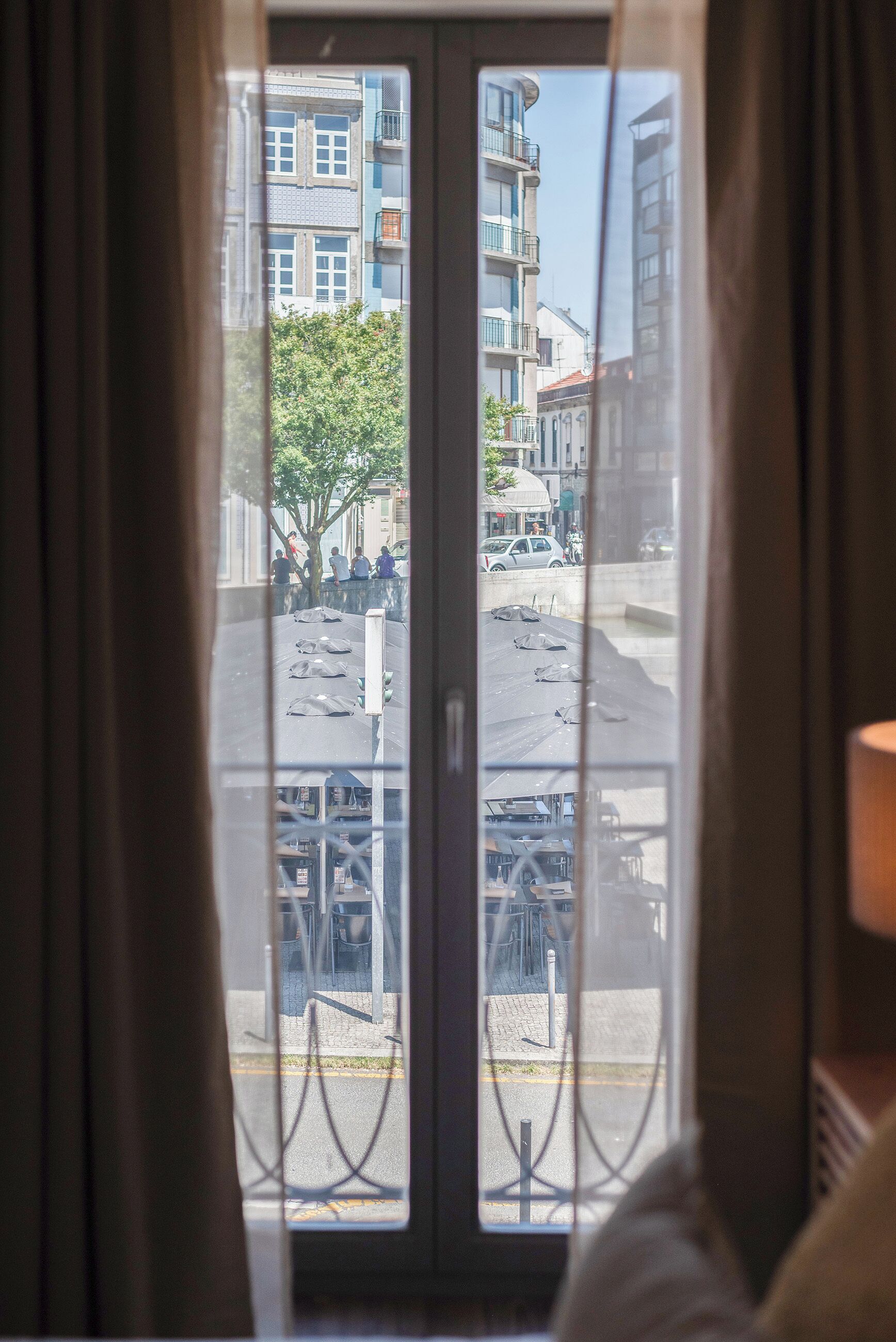 A view through a window with curtains, featuring a street scene with buildings and parked cars.