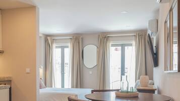 A hotel room with a bed, a dining table with chairs, a kitchen area, and a large window with curtains.