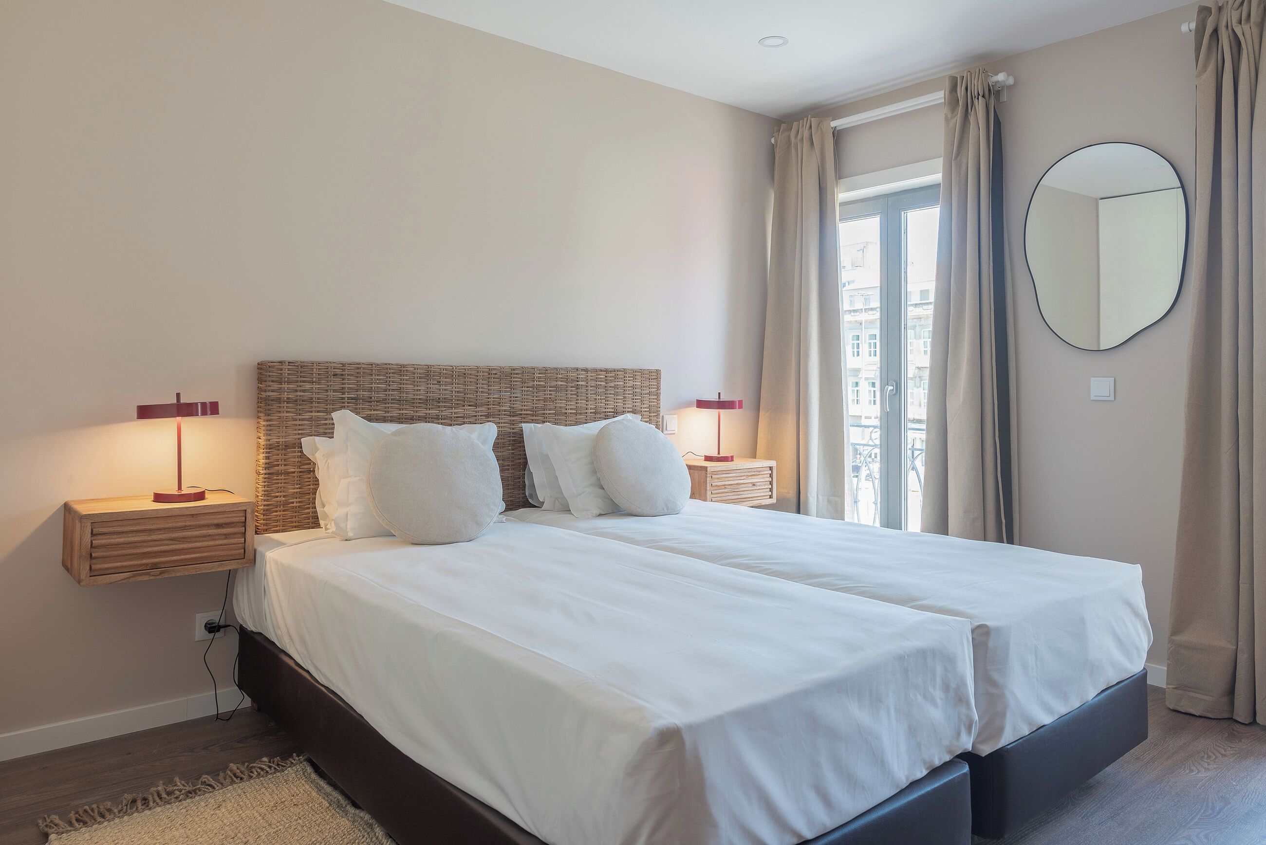 Un lit double avec du linge de lit blanc, deux tables de chevet avec des lampes rouges, une tête de lit en osier et un grand miroir au mur.