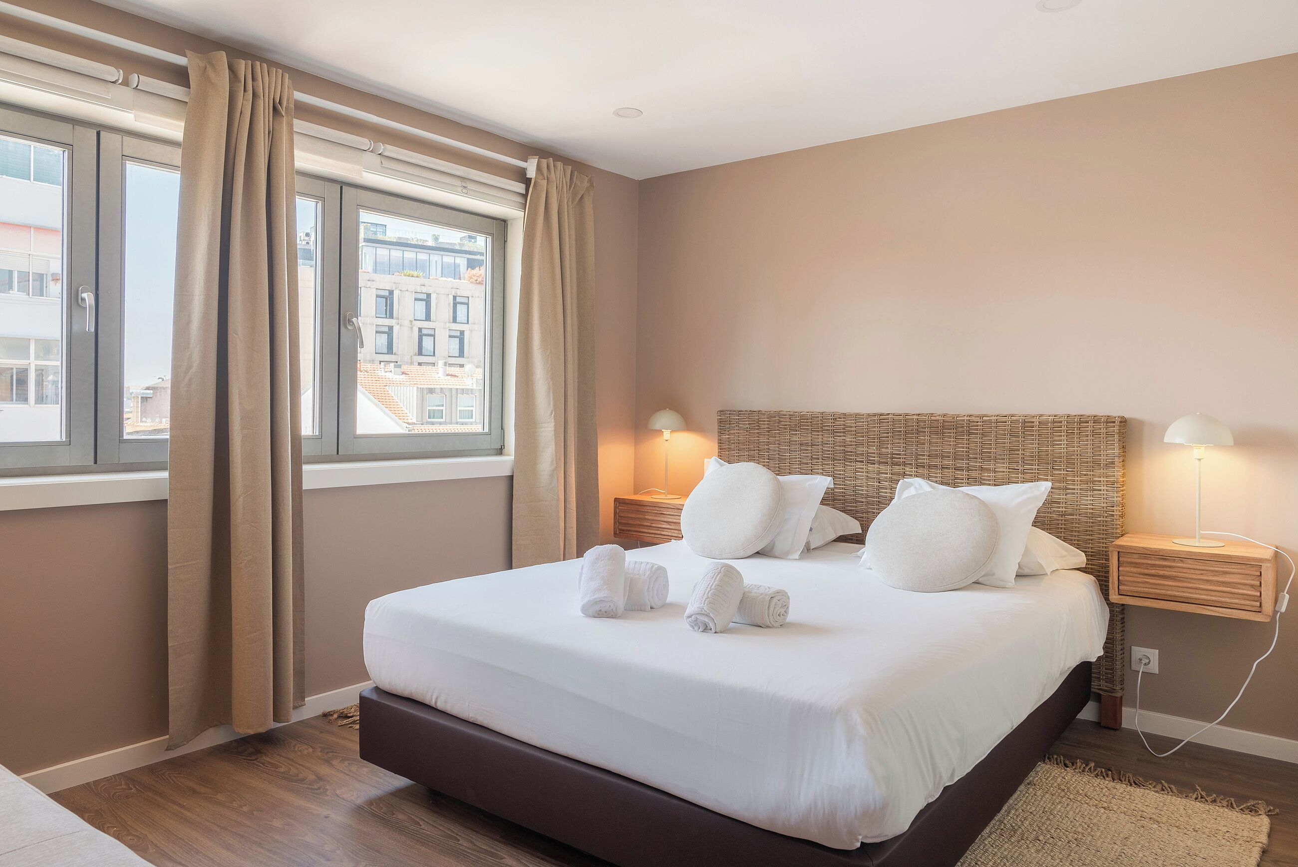 Een net opgemaakt bed met witte lakens en opgerolde handdoeken, een rieten hoofdbord, nachtlampjes en een uitzicht op gebouwen door grote ramen met beige gordijnen.