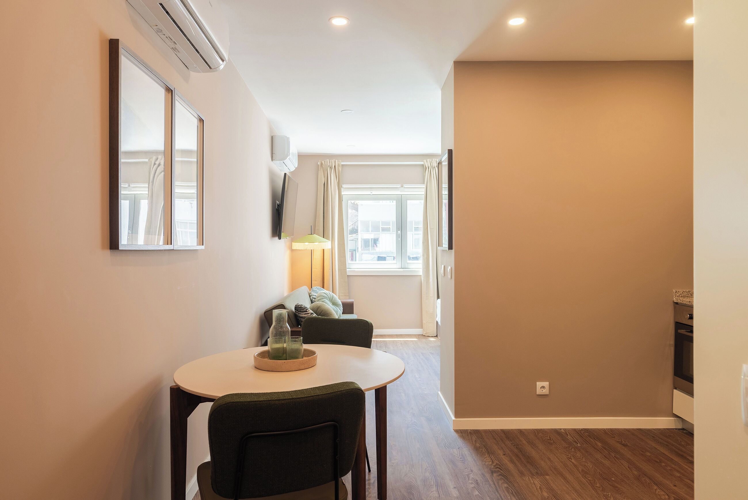 A modern living room with a dining area, a flat-screen TV mounted on the wall, and a window with curtains.