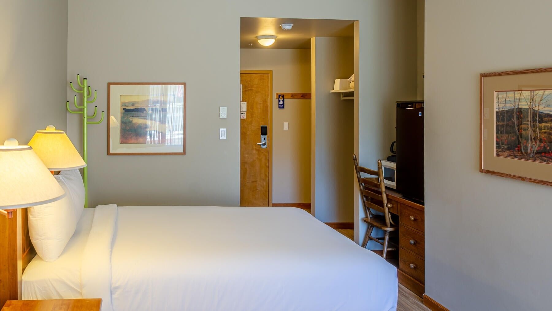 Quarto de hotel com cama, escrivaninha, cadeira, luminária e um quadro na parede.