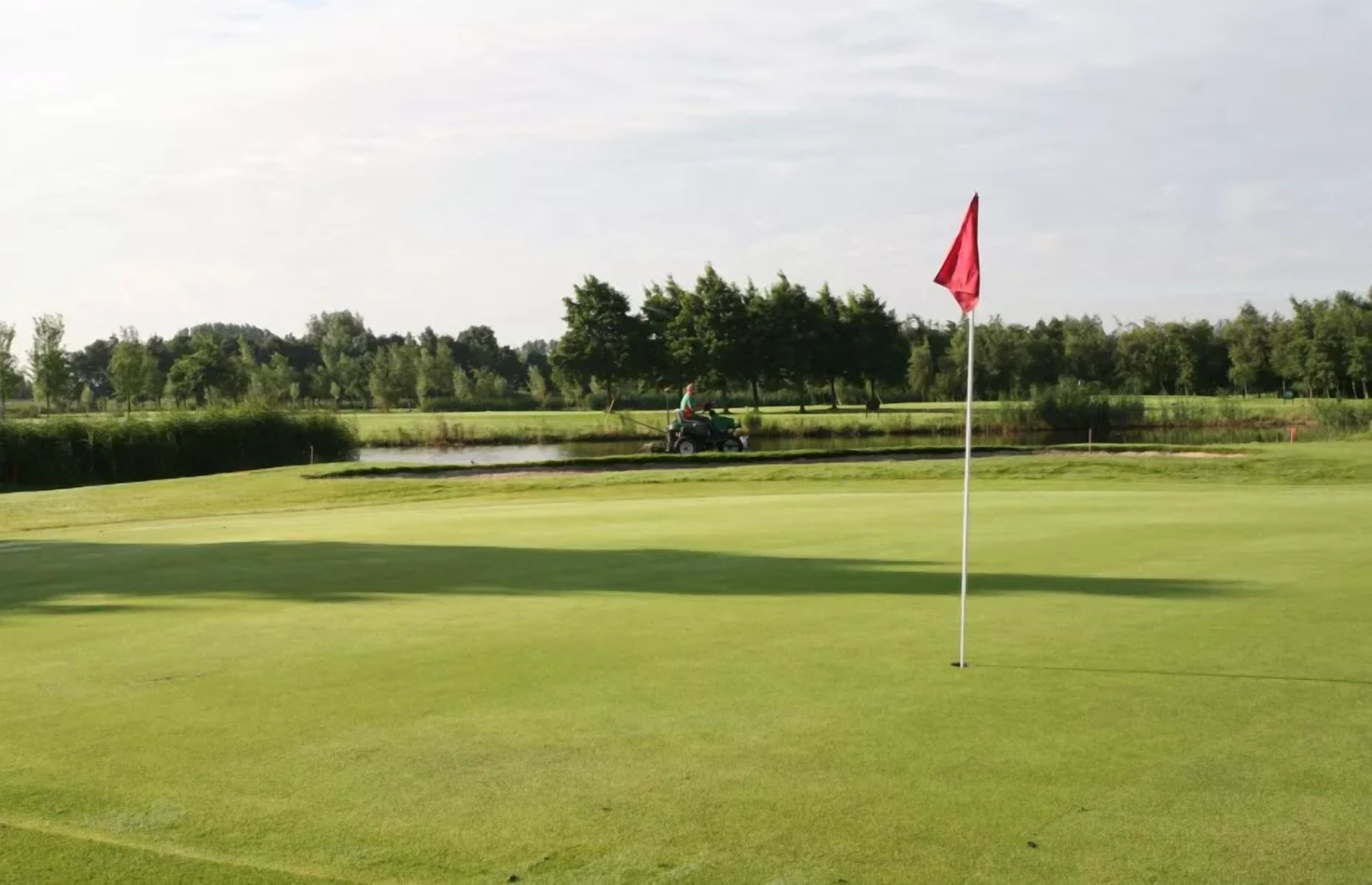 A golf course with a flag, a pond, and a tractor in the background.