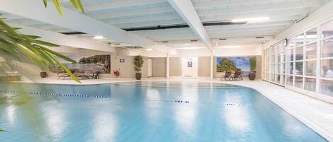 An indoor swimming pool with clear blue water, surrounded by a tiled floor and walls. There are lounge chairs and potted plants around the pool area.