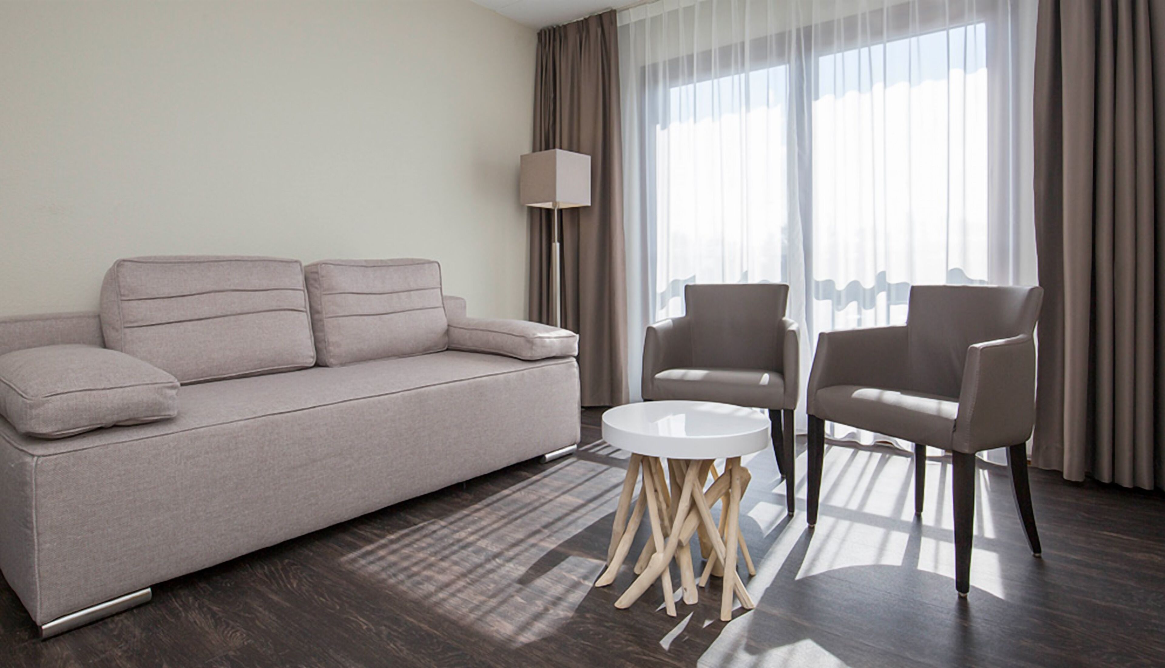 A modern hotel room with a grey sofa, two armchairs, a small white table, and a floor lamp.