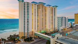Un hôtel de grande hauteur doté de nombreux balcons, offrant une vue sur la plage et relié à un pont piétonnier.