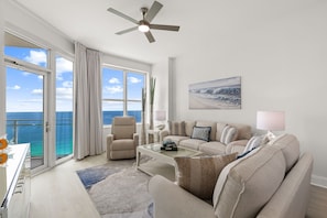 A spacious living room with a large window, a ceiling fan, a sofa, a coffee table, and a painting on the wall. - Aqua 2310 PCB at its Finest/Oceanfront/ High Clean Standard/Wifi (Panama City Beach)