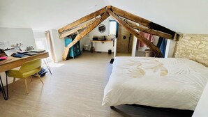 A bedroom with a sloped ceiling, a bed with a white bedspread, a desk with a chair, and a bathroom visible in the background.