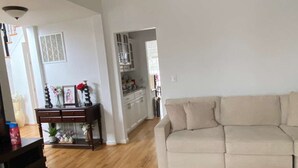 Una sala de estar con un sofá color beige, piso de madera, una mesita de noche de madera oscura y un aparador de vidrio con objetos decorativos.