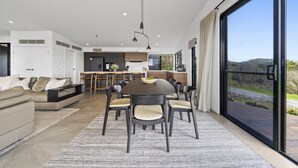 A modern living room with a sofa, a dining table with chairs, and a kitchen in the background. - Scenic House With Private Yard & BBQ Area (Springbrook)