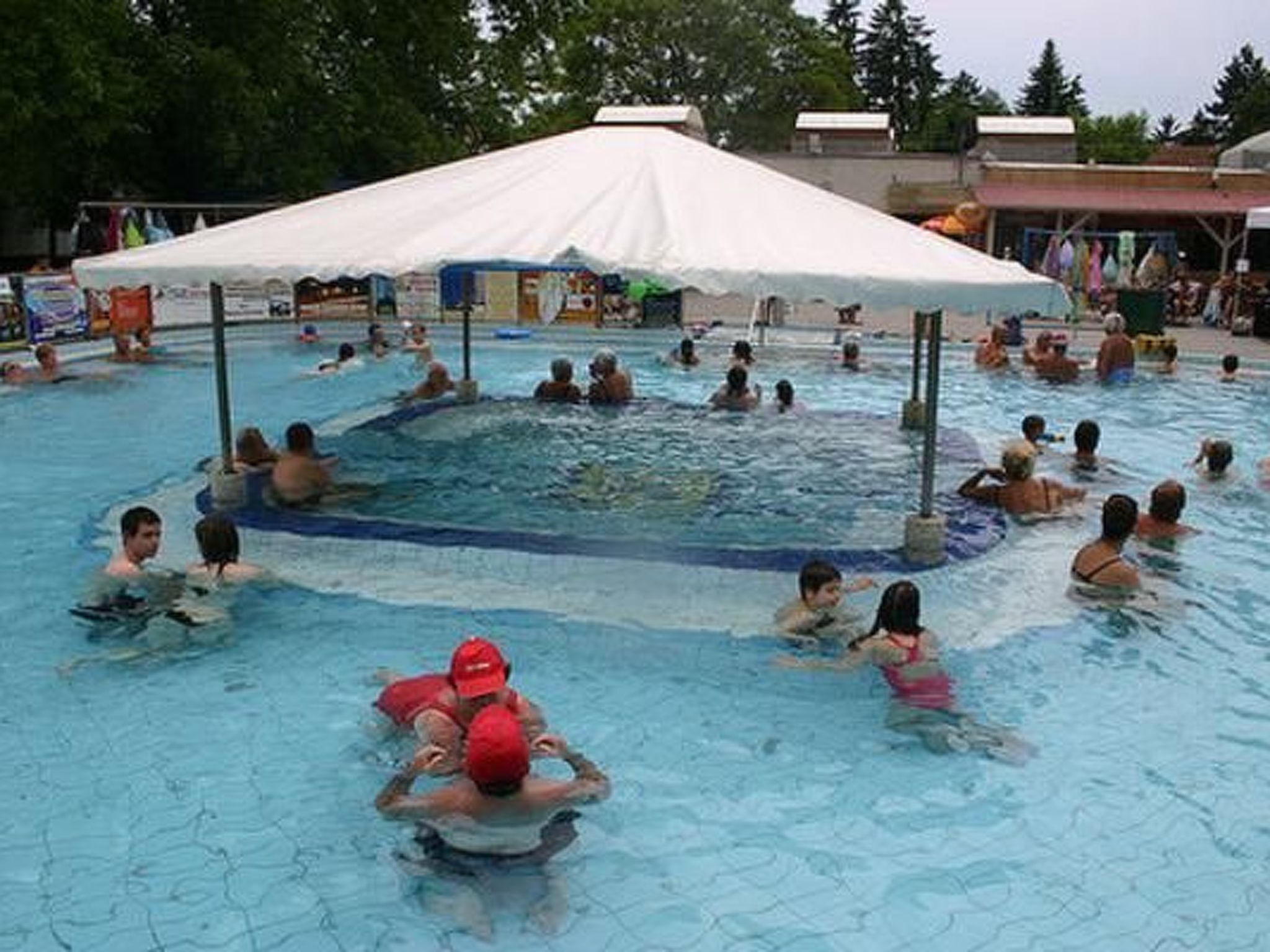 Una piscina con gente nadando, una zona cubierta y un edificio al fondo.