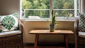Ein gemütliches Zimmer mit Blick auf Bäume durch ein Fenster, ein Korbssofa mit gemusterten Kissen und ein kleiner Holztisch mit einer Topfpflanze.