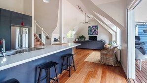 Eine moderne Küche mit einer großen Kochinsel und Barhocker, eine Treppe zu einem Erkerbereich und ein gemütlicher Wohnbereich mit Sofa und Fernseher.
