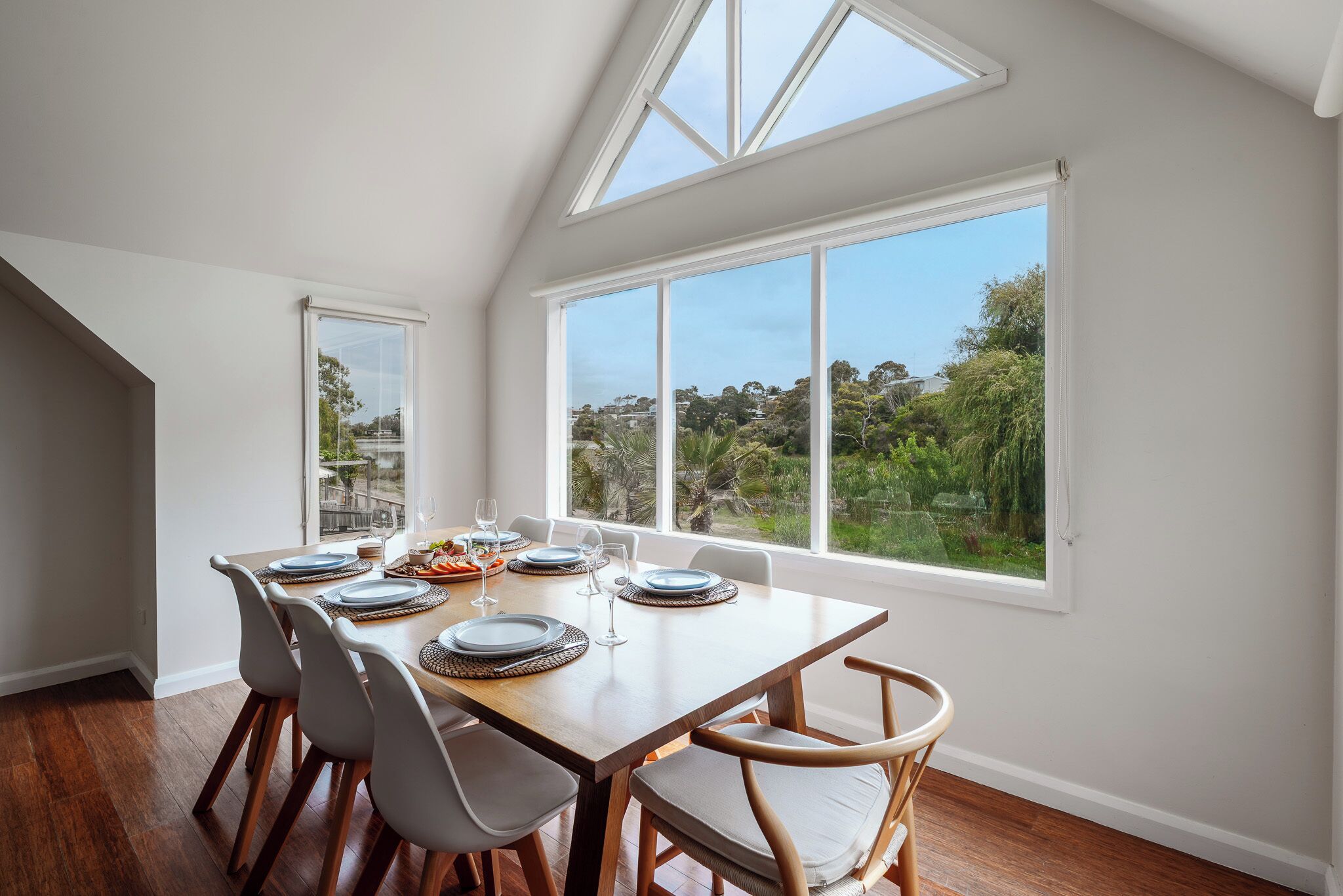 A dining area with a wooden table set for four, large windows offering a view of greenery and buildings, and a sloped ceiling with skylights.