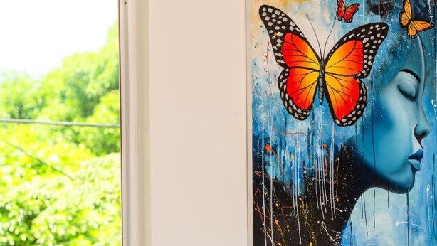 Quarto com janela, pintura de borboleta e rosto, e cadeira de vime.