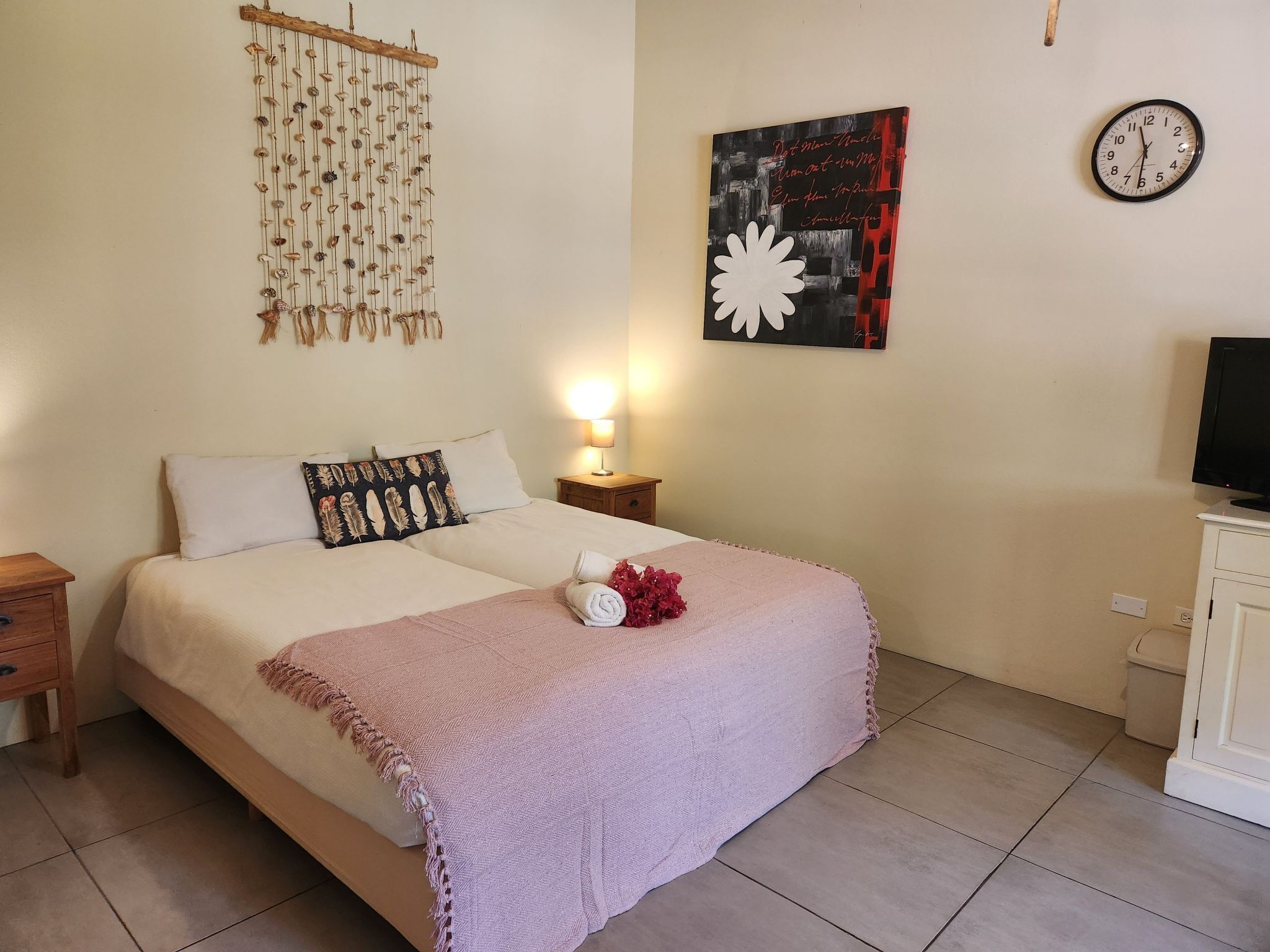 Ein Doppelbett mit einer rosa Decke, Wanddekoration mit Muscheln, eine Uhr und ein Gemälde.
