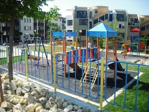 A playground with various play structures, surrounded by a metal fence, and residential buildings in the background. - Apartment Vega by Interhome (St. Vlas/Sveti Vlas)