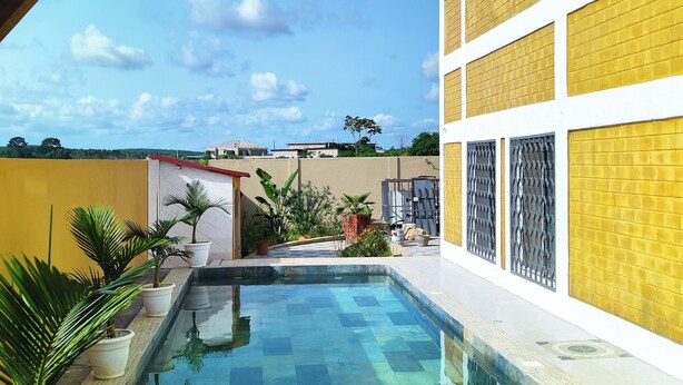 A swimming pool with a clear blue water surface, surrounded by potted plants and a yellow building with white windows. - Villa Macarena (Bonoua)