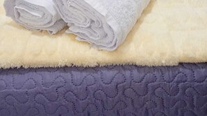 Ein ordentlich bezogenes Bett mit einem gefalteten weißen Handtuch und einer beigen Tagesdecke auf einem strukturierten violetten Bettbezug.