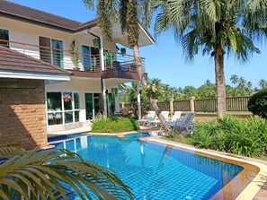 A modern two-story house with a swimming pool, surrounded by palm trees and lush greenery.