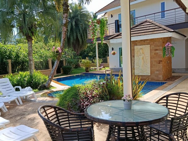 A pool area with lounge chairs, a glass table, and a house with a balcony.