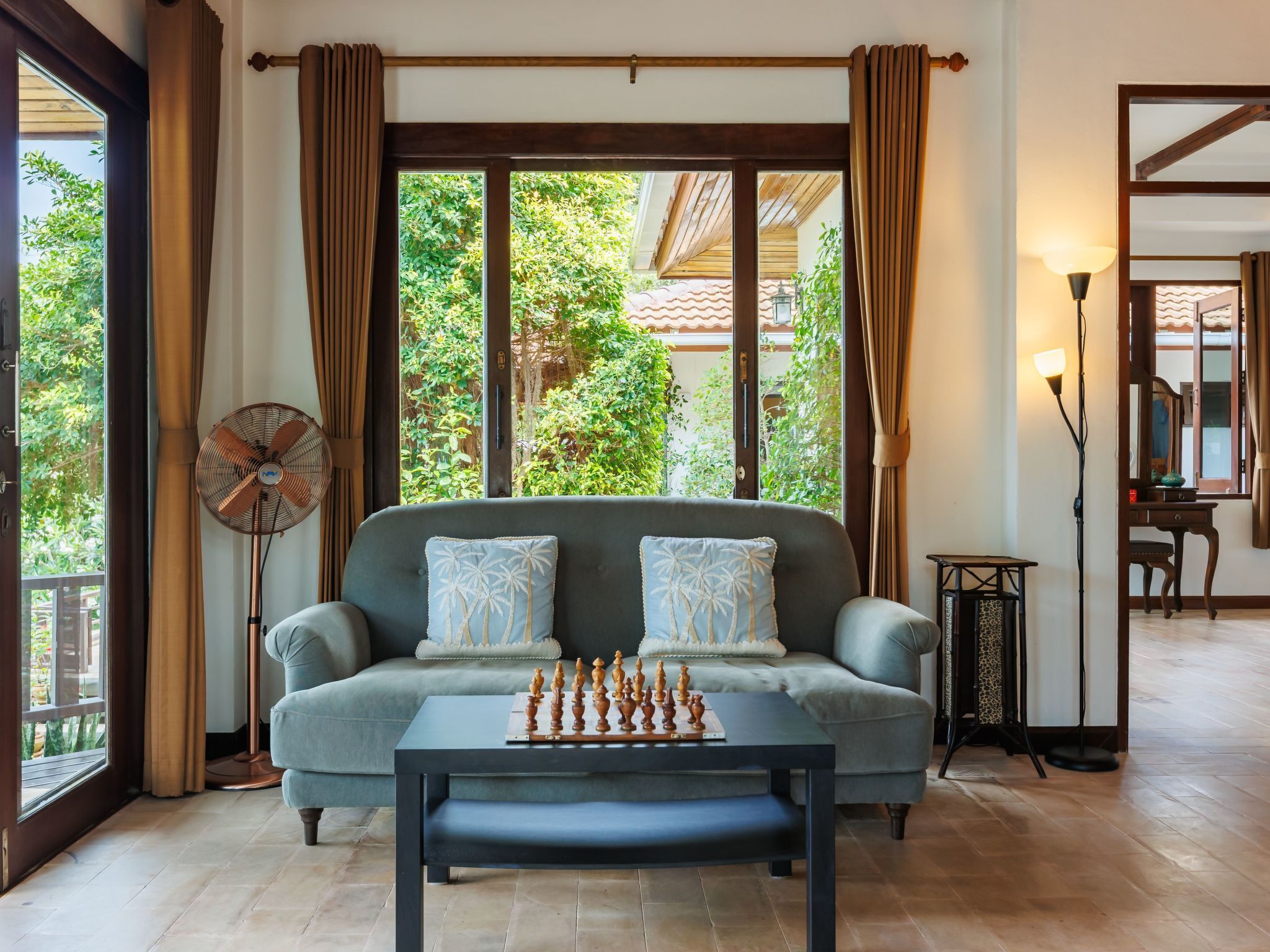 Un salon comprenant un canapé, une table d’échecs, un ventilateur sur pied et une vue sur la verdure grâce à de grandes fenêtres.