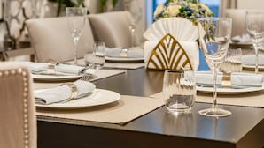En matplats med ett bord dukat för fyra personer, inklusive tallrikar, bestick och glas. På bordet står en dekorativ vas med blommor, och i bakgrunden finns en växt.