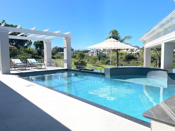 A swimming pool with white lounge chairs and a pergola, surrounded by greenery and houses in the background. - in Bos En Dal mit Großer Terrasse by Interhome (George)