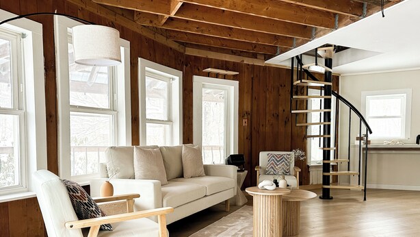 Un vaste salon doté d’un plafond en bois, d’un canapé blanc, d’une chaise en bois, d’une table basse ronde en bois et d’un escalier en colimaçon.