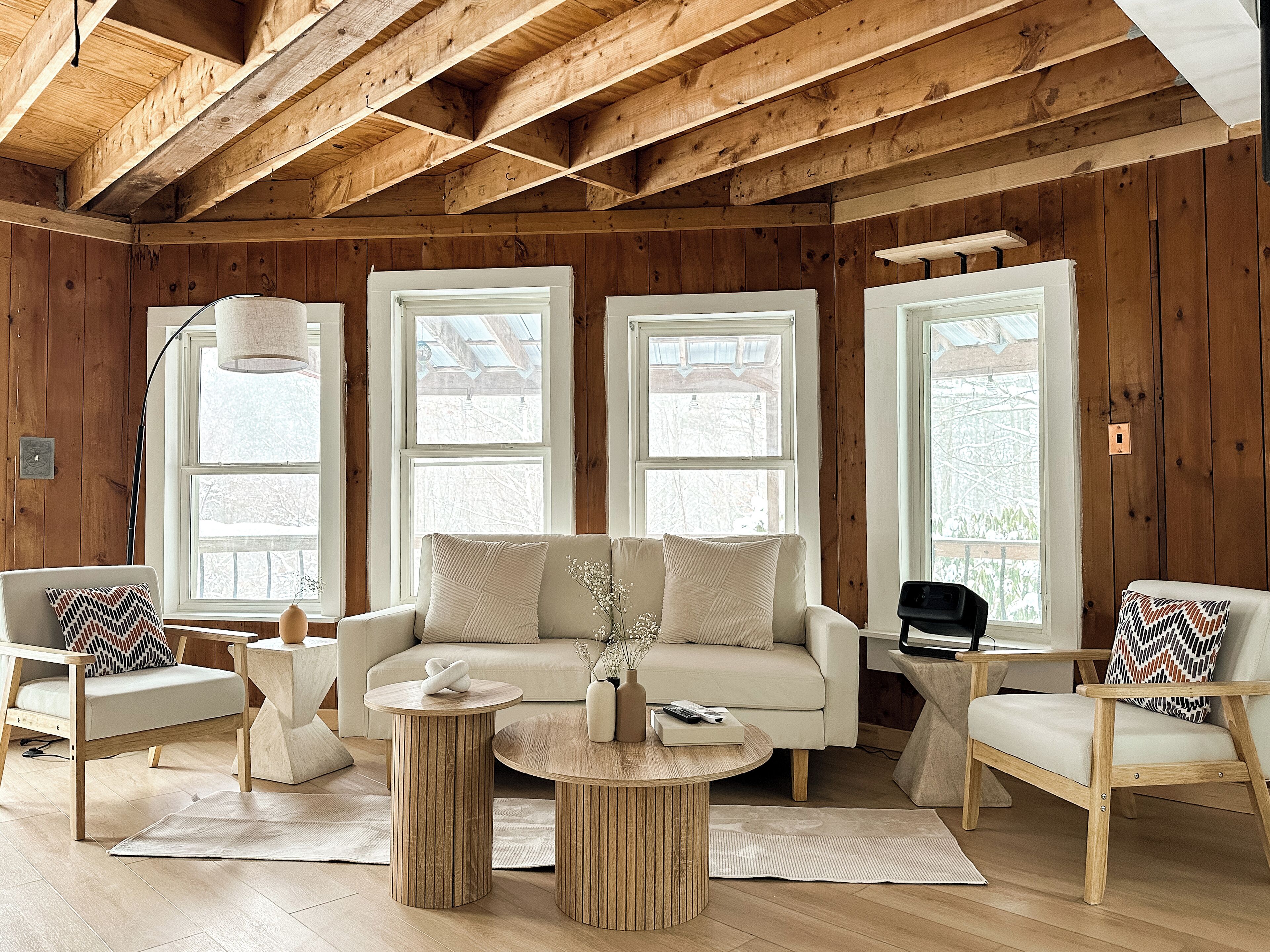 Ett mysigt vardagsrum med ett träpanelat tak, stora fönster, en beige soffa, två fåtöljer och ett runt soffbord.
