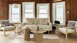 Un salon chaleureux avec un plafond en bois, de grandes fenêtres, un canapé beige, deux fauteuils et une table basse ronde.