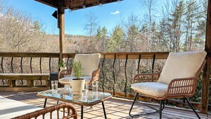 Une terrasse en bois avec des chaises en osier et une table en verre, donnant sur une zone boisée.