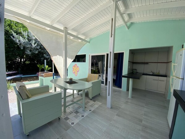 Un espacio al aire libre cubierto con mesa y sillas, un sofá y una zona de cocina con armarios y un refrigerador.