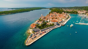 Vue aérienne d’une ville côtière, avec une imposante structure fortifiée, un port de plaisance rempli de bateaux et une route menant à la ville.