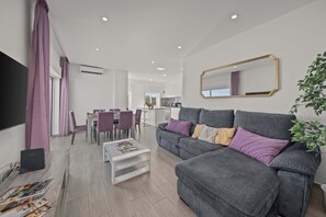 A modern living room with a grey sofa, white coffee table, and purple cushions. A dining area with a table and chairs is visible in the background. - Elegant Flat with Balcony in Lagos by HostWise (Lagos)