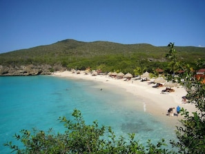 Una playa con aguas turquesas cristalinas, cabañas con techo de paja y un paisaje montañoso al fondo.