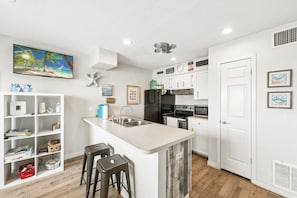 A kitchen with a central island, white cabinetry, a refrigerator, and a microwave. There are bar stools, a shelf with items, and a painting on the wall. - Waterfront Island Escape | Balcony, Boat Slips & Pool (Corpus Christi)