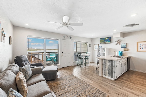 A spacious living area with a ceiling fan, a dining table with chairs, a kitchen island, and a view through large windows. - Waterfront Island Escape | Balcony, Boat Slips & Pool (Corpus Christi)